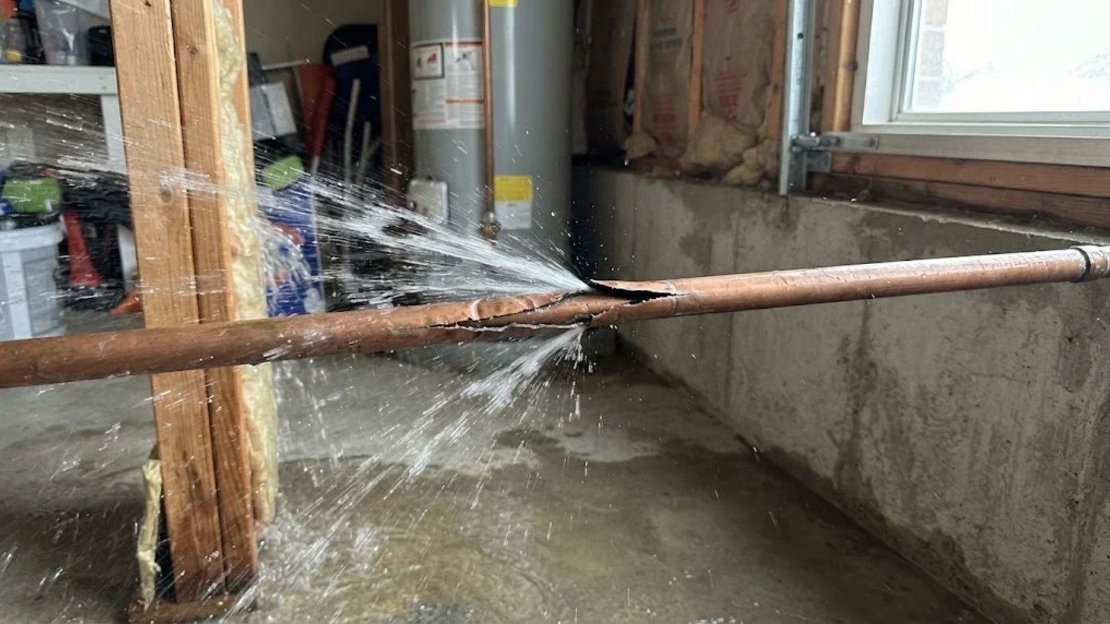 A burst copper pipe spraying water along a basement wall and over the floor.