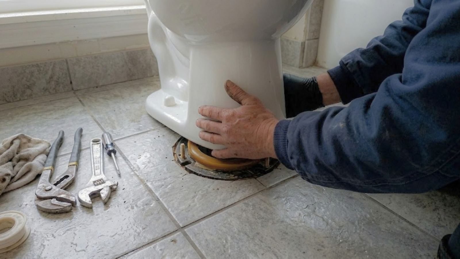 A plumber pressing a toilet onto its floor flange with tools laid out on the tile.